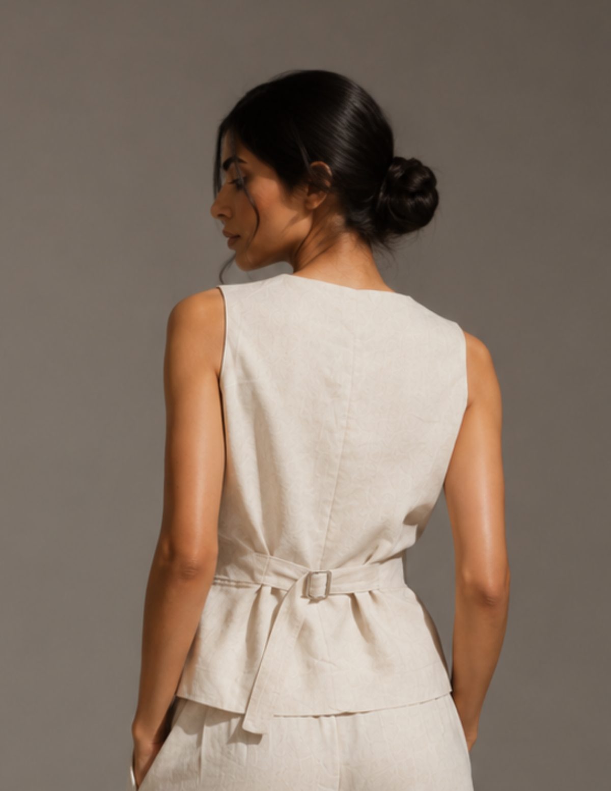 A woman shown from the back wearing a sleeveless light beige linen belted top and matching bottoms against a plain gray studio background.