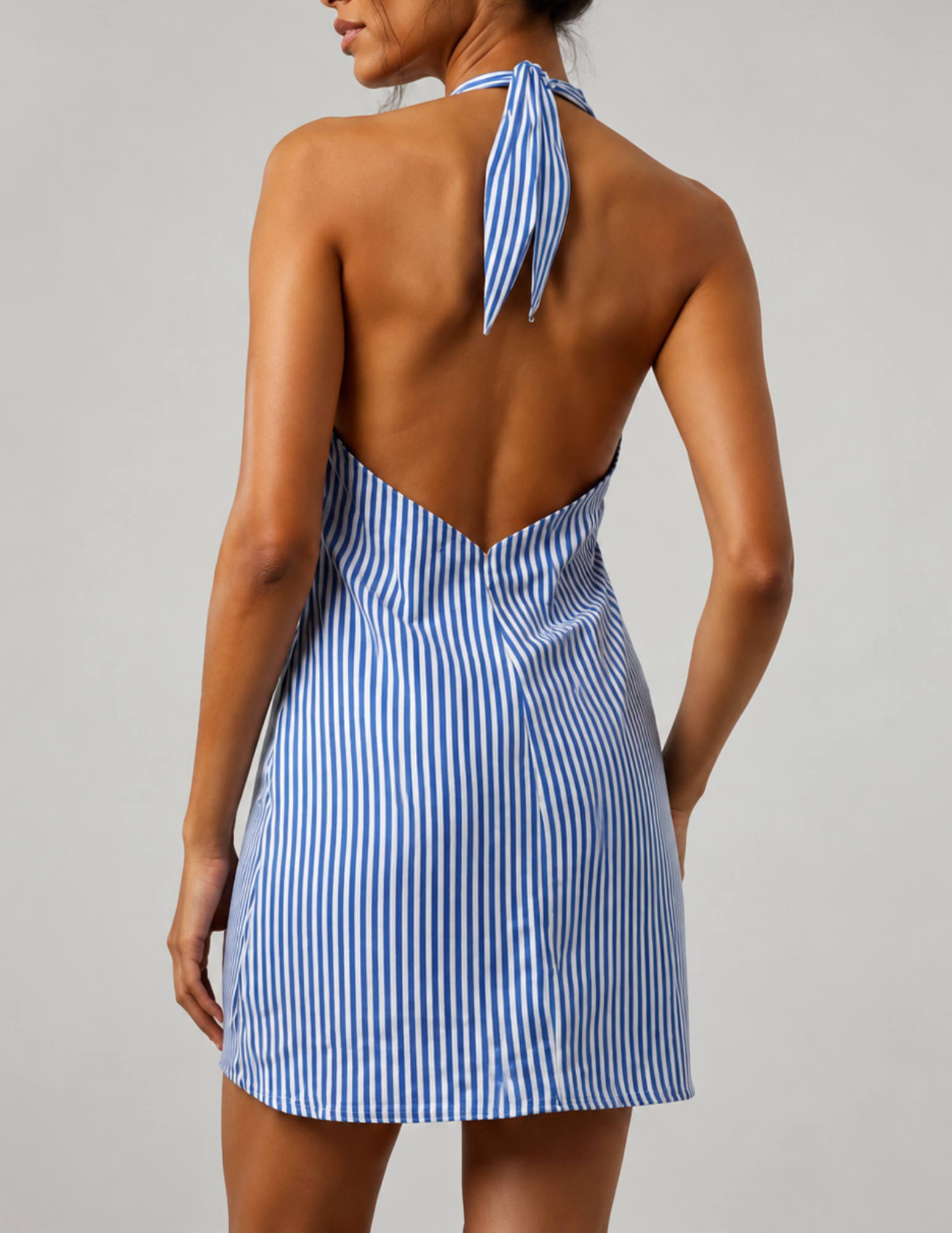 A woman shown from the back wearing a blue and white vertically striped halter mini dress with an open back, photographed against a plain light gray studio background.
