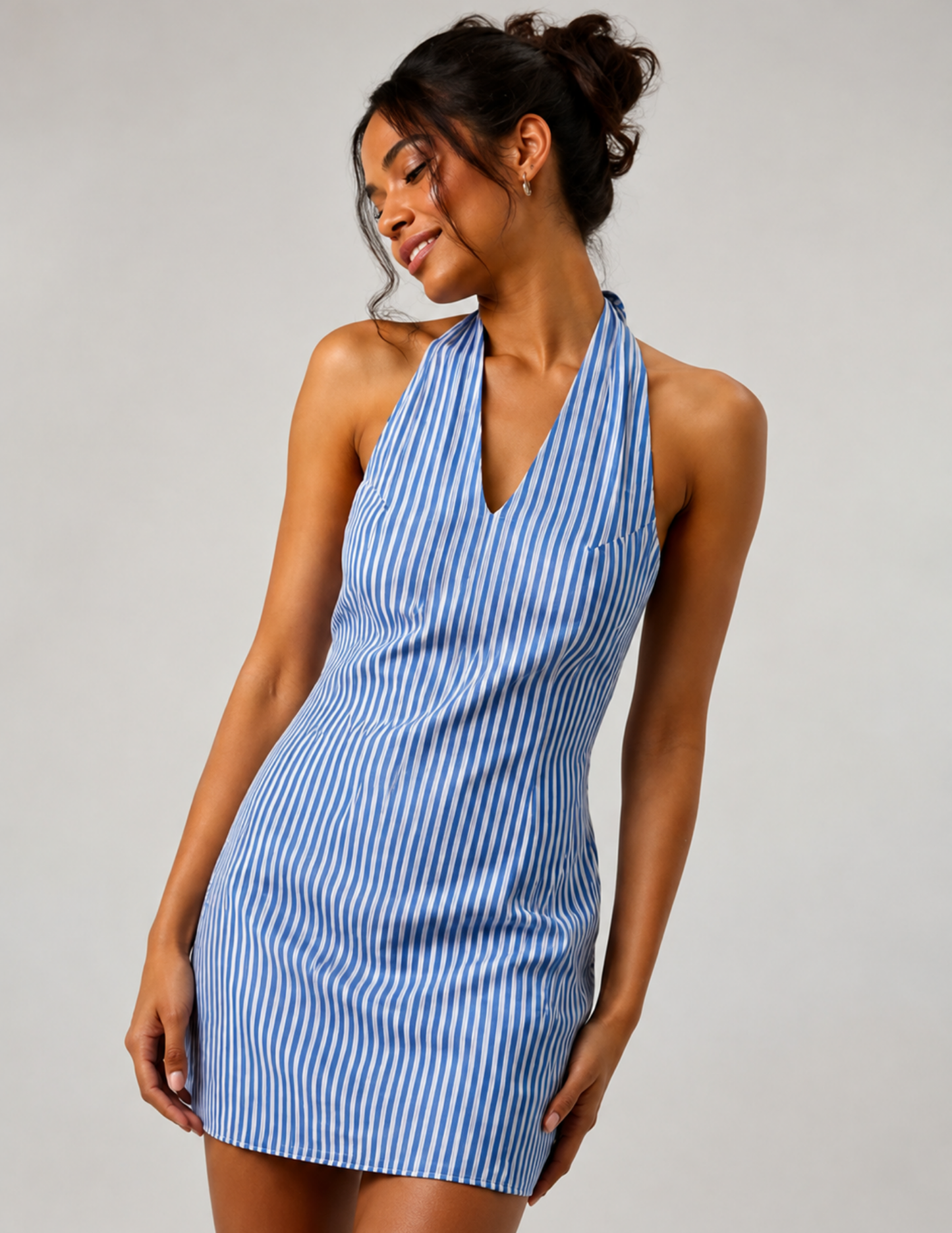 A woman modeling a sleeveless halter-neck mini dress with vertical blue and white stripes, posed against a neutral light gray studio background.