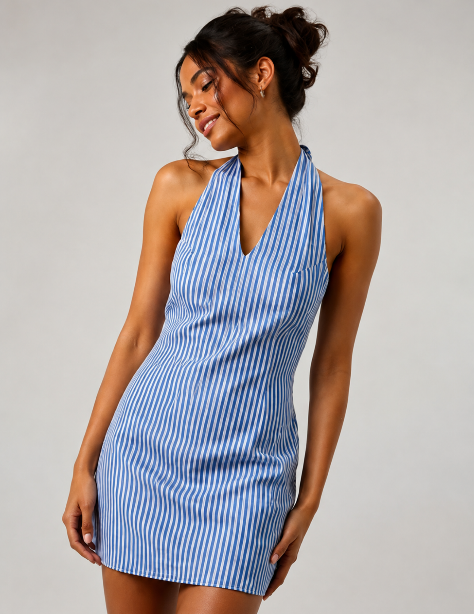 A woman modeling a sleeveless halter-neck mini dress with vertical blue and white stripes, posed against a neutral light gray studio background.