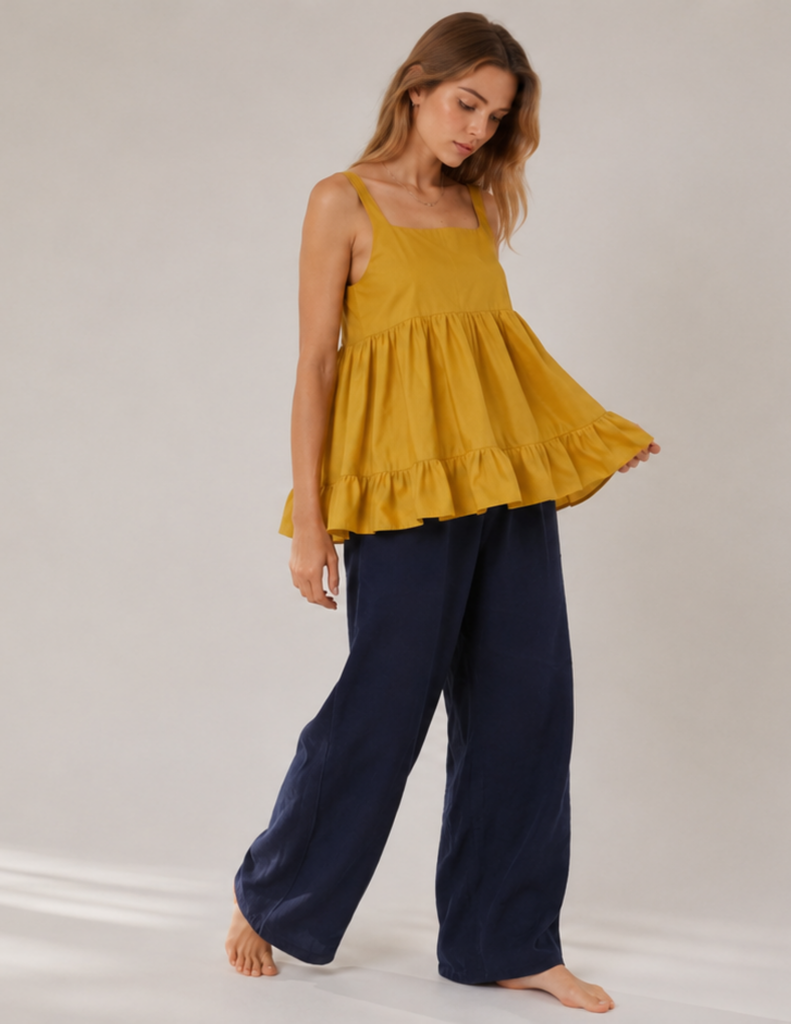 A woman modeling a sleeveless mustard yellow peplum top paired with dark navy wide-leg pants, standing against a light gray studio background.