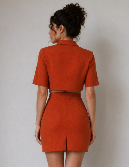 A woman shown from the back wearing a matching rust-orange cropped blazer and high-waisted mini skirt set against a plain light gray studio background.