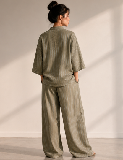 A woman stands with her back to the camera wearing a matching two-piece lounge set in a muted sage green color with thin vertical stripes. The set includes a short-sleeve top and wide-leg pants, photographed in a minimalist room with warm natural light casting soft shadows on the wall.