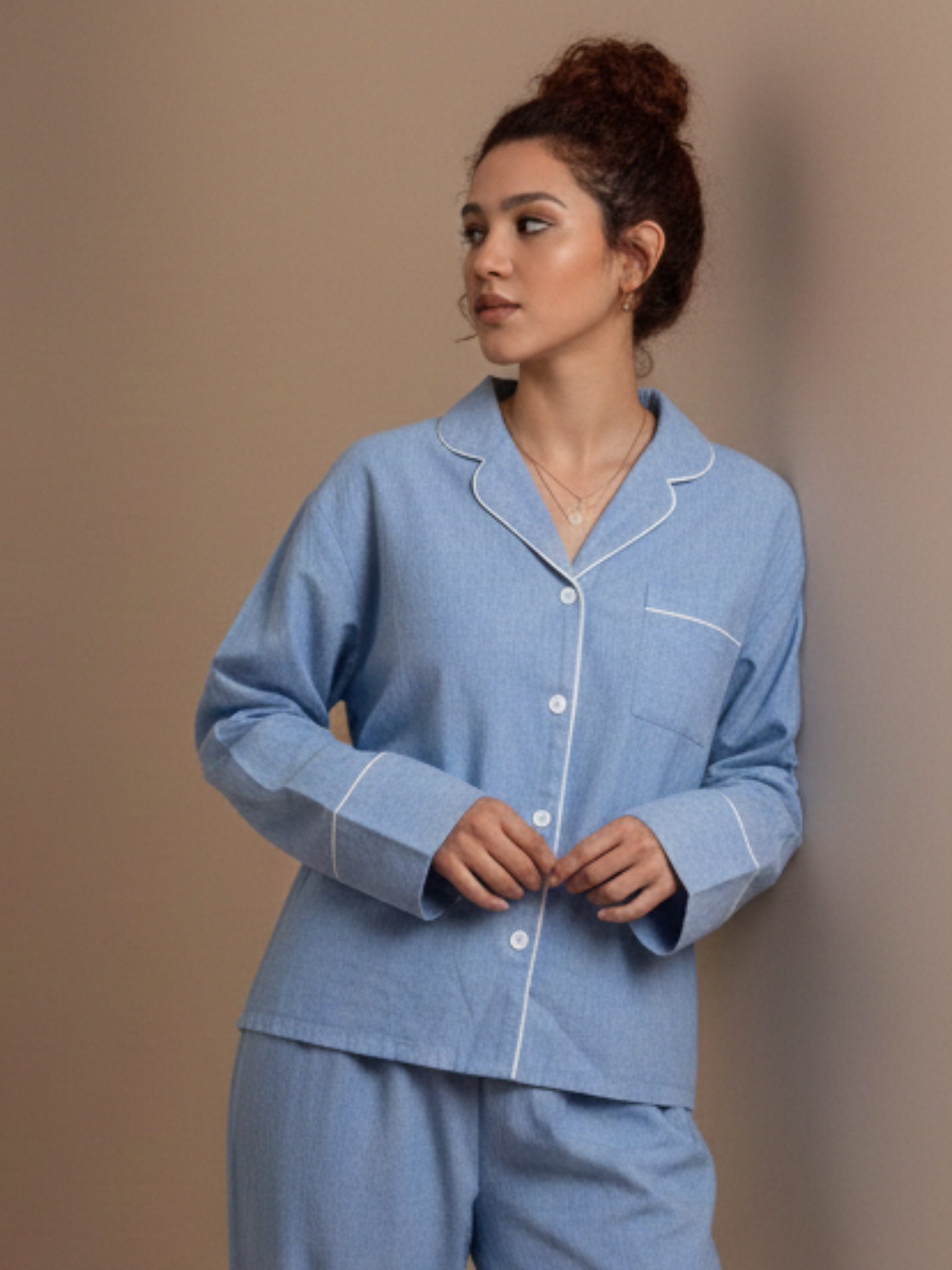 A woman wearing a light blue pajama set with white piping details, standing against a solid beige background.