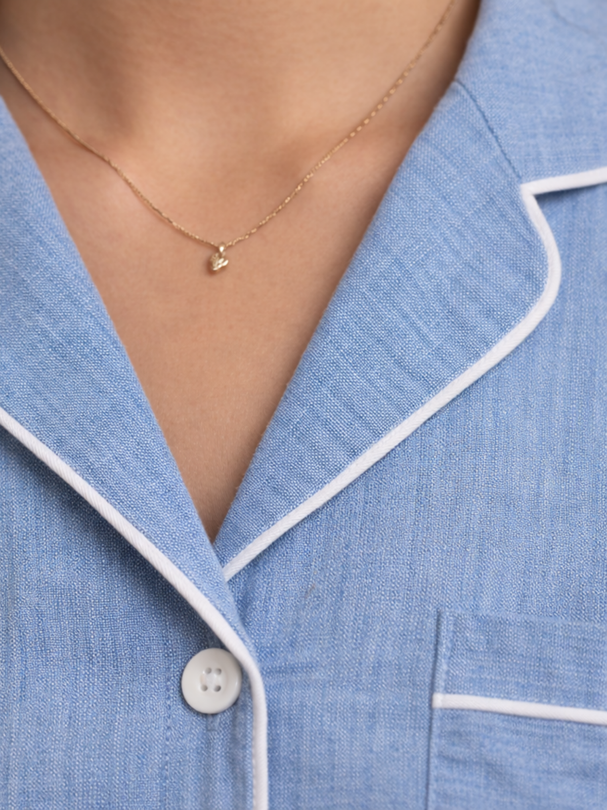 Close-up of a person wearing a light blue pajama top with white piping trim and a delicate gold necklace.