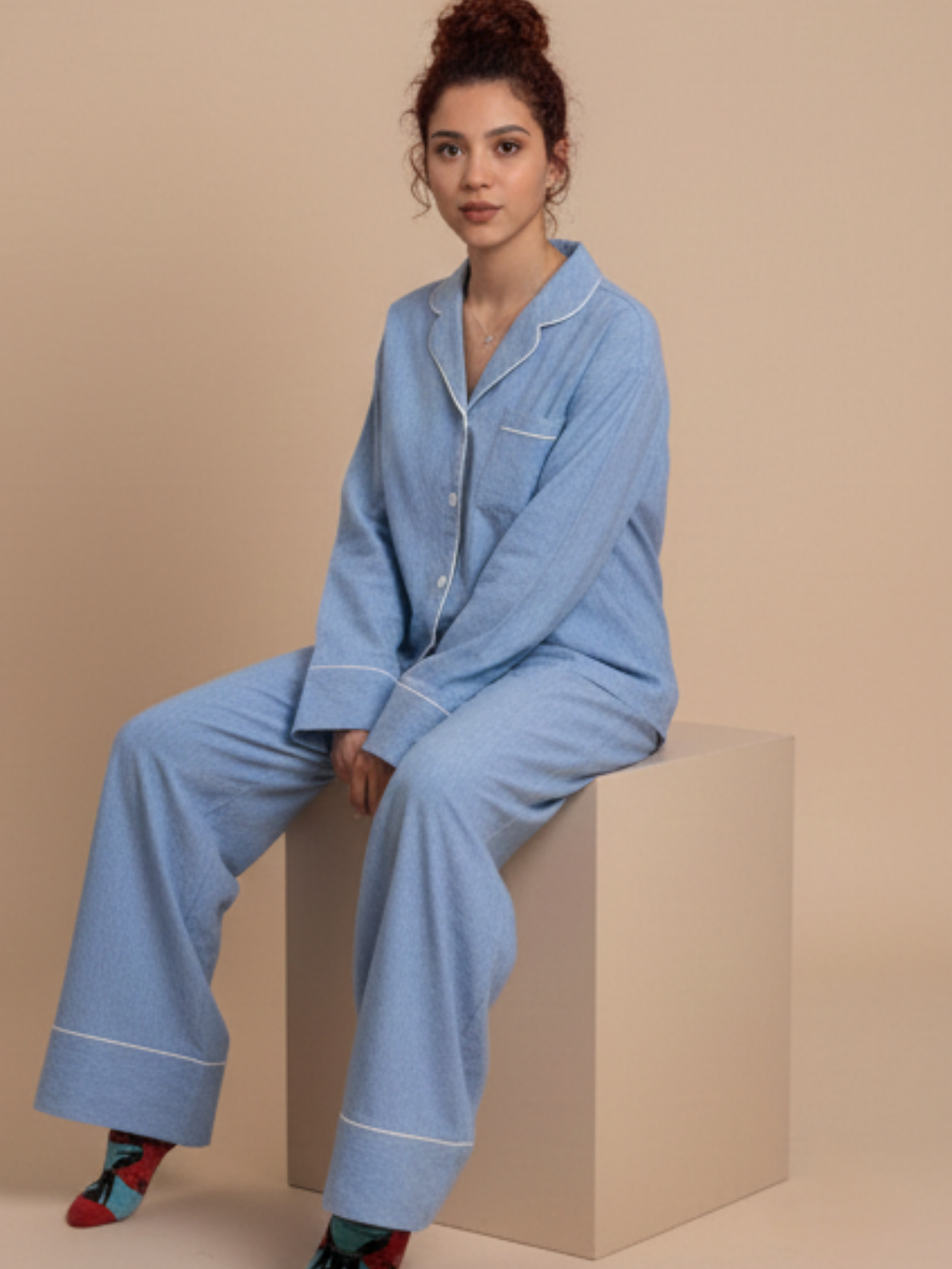 A woman wearing a light blue pajama set with white piping sits on a beige block against a solid beige background.
