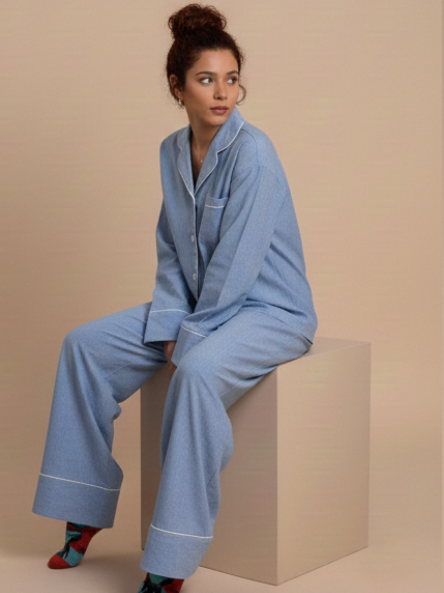 A woman wearing a light blue pajama set with white piping, sitting on a beige block against a solid beige background.