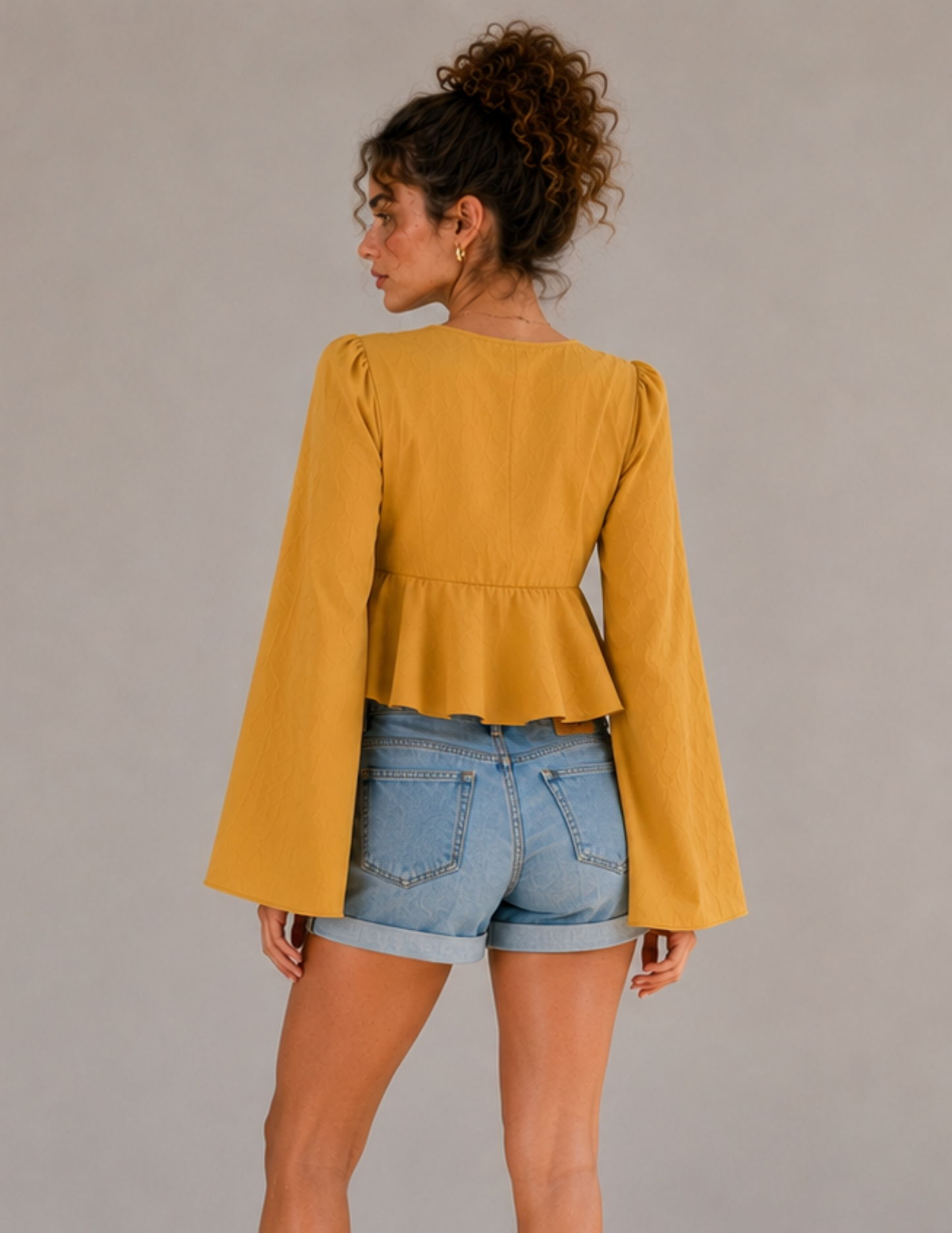 MARBELLA peplum top sewing pattern shown from the back, featuring long flared sleeves, gathered sleeve caps, fitted bodice and gathered peplum hem in mustard yellow fabric.