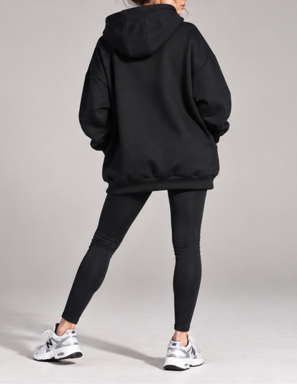 A person shown from the back wearing an oversized black hoodie, black leggings, and white New Balance sneakers against a light gray studio background.
Back view of the Melani women's 
oversized hoodie showing the 
relaxed silhouette, spacious hood, 
dropped shoulders, voluminous 
sleeves and ribbed bottom band 
extending below the hips.