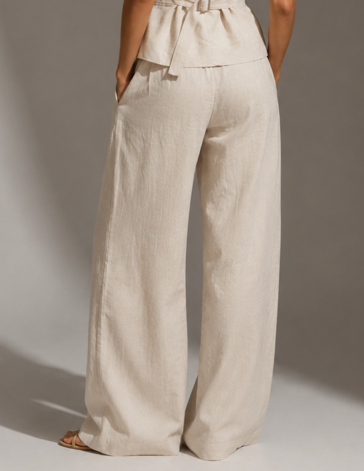 A person shown from the back wearing high-waisted, wide-leg pants in a light warm off-white or cream linen-like fabric, photographed against a plain gray studio background.
