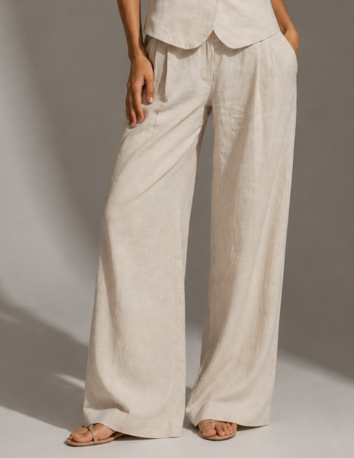 A person wearing loose, wide-leg linen pants in a light oatmeal beige color, paired with matching top and flat sandals, photographed against a neutral studio backdrop.