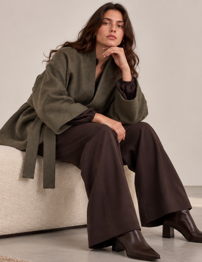 Woman wearing a relaxed oversized wrap coat with a shawl collar, wide kimono sleeves, waist belt, and patch pocket in textured olive green fabric.