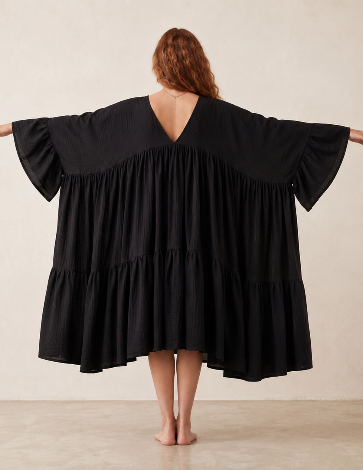 A woman viewed from behind wearing a loose black tiered tunic dress with wide flutter sleeves and a V-neckline, standing barefoot against a plain beige wall.