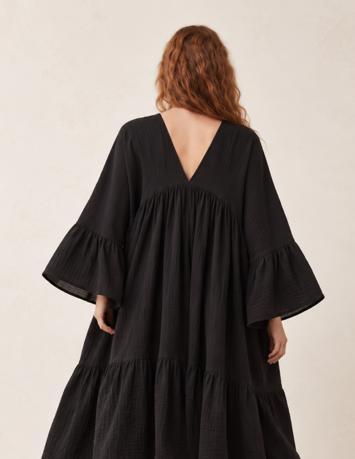 A woman viewed from the back wearing a loose black tiered dress with wide flared sleeves and a deep V-neckline, photographed against a plain light beige studio background.