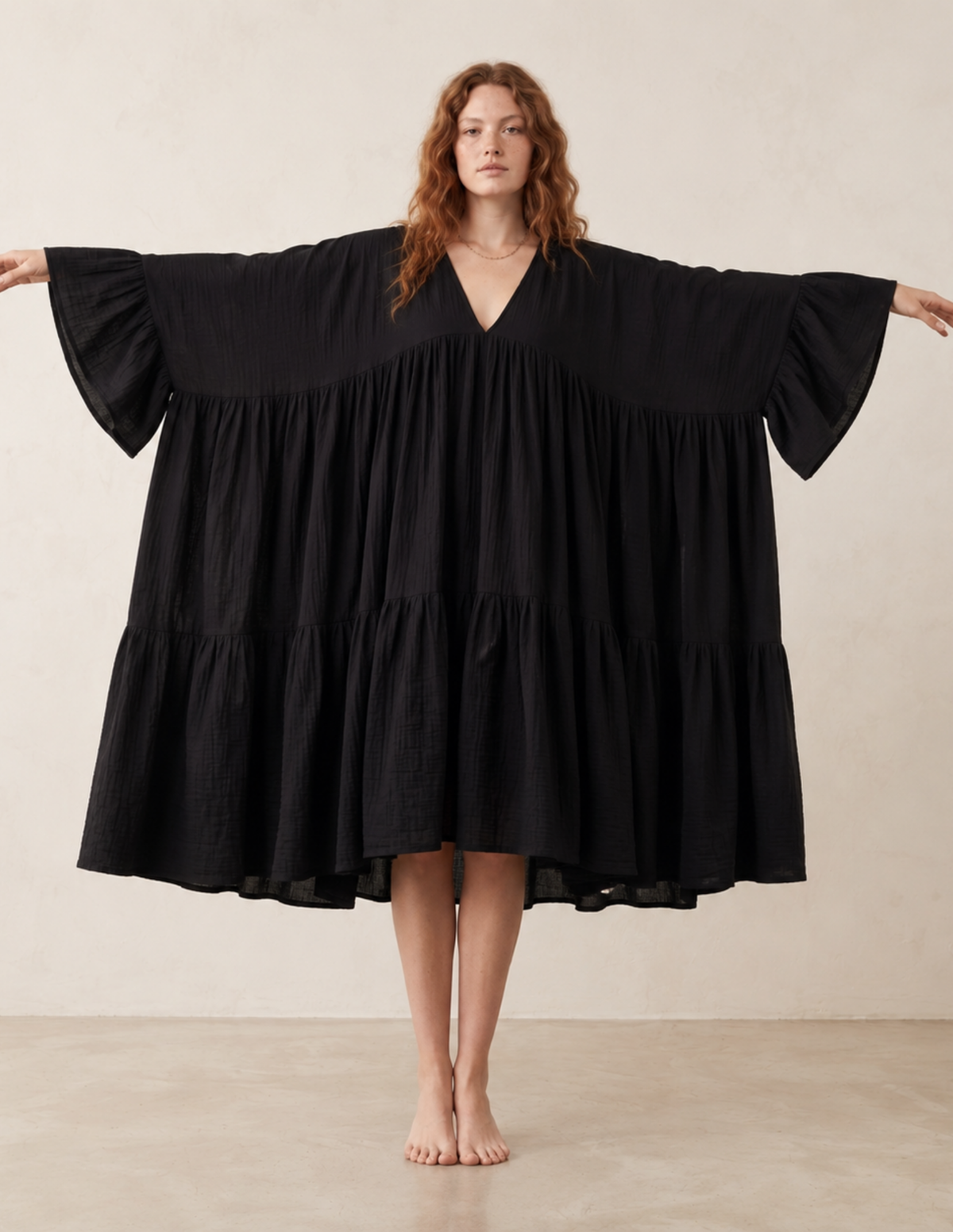 A woman stands barefoot with her arms outstretched, wearing a loose black tiered dress with wide flared sleeves against a plain beige studio background.