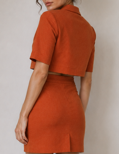 A woman wearing a matching burnt orange two-piece set consisting of a short-sleeve cropped collared top and a high-waisted mini skirt, shown from a rear three-quarter angle against a neutral light gray background.
