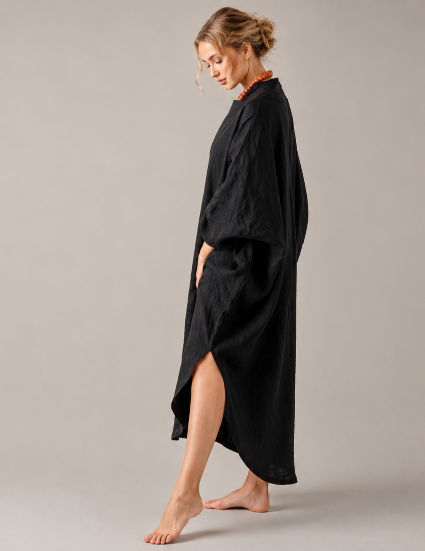 A woman modeling a loose, black, high-slit kaftan-style robe against a solid light gray studio background.